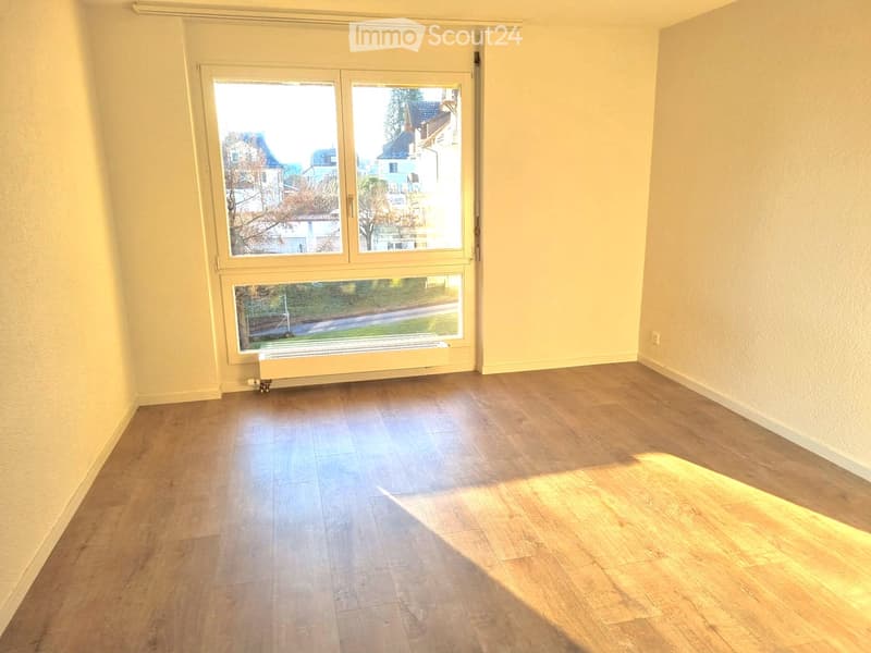 Empty room with wooden flooring and large windows offering a view of the street and surrounding buildings.