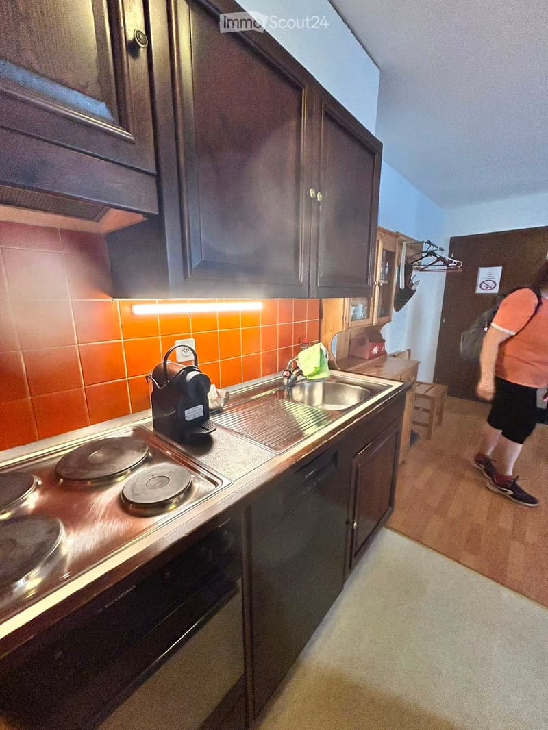 Compact kitchen with wooden cabinets, a stove, sink, and dishwasher. The countertop features a coffee maker and a kettle. The backsplash is orange tiled.