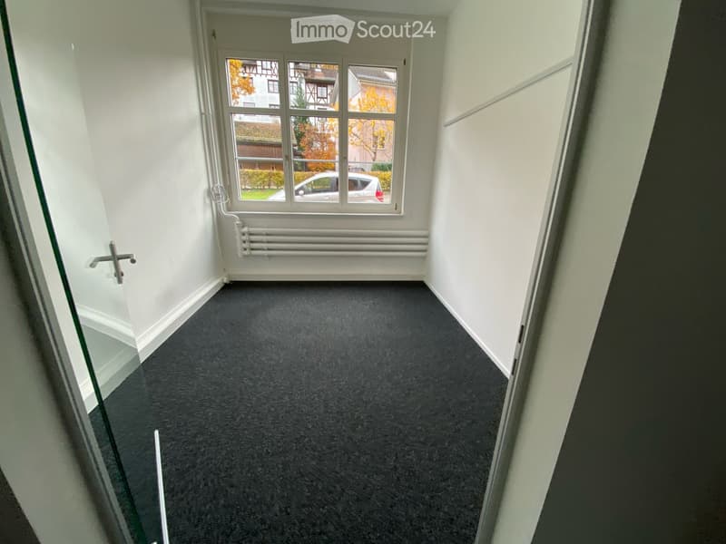 Empty room with white walls, dark grey carpet, large window with view of the street, radiator, and door handle visible.
