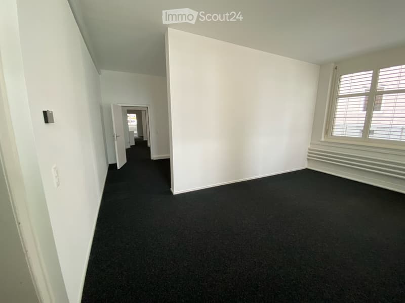 Empty room with white walls, black carpet, and a window with blinds. No furniture or equipment is present.