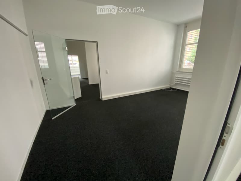 Empty room with white walls, black carpet, and a glass door leading to another space.