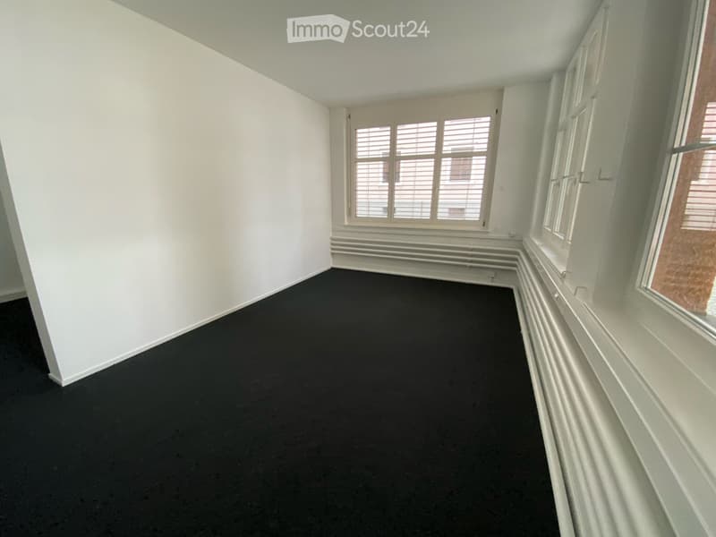 Empty room with white walls, black carpet, and large windows with blinds.