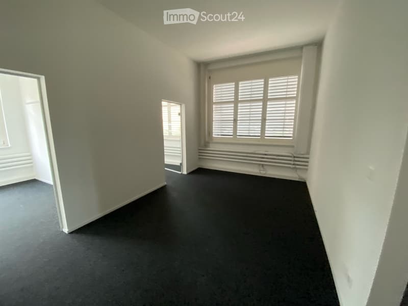 Empty room with light grey walls, dark grey carpeted floor, large window with white shutters, radiator below window, and an open doorway leading to another space.