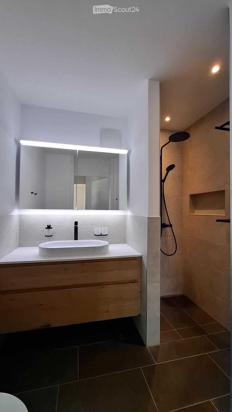 Modern bathroom with a white sink, wooden cabinet, large mirror with LED lighting, and a handheld showerhead.