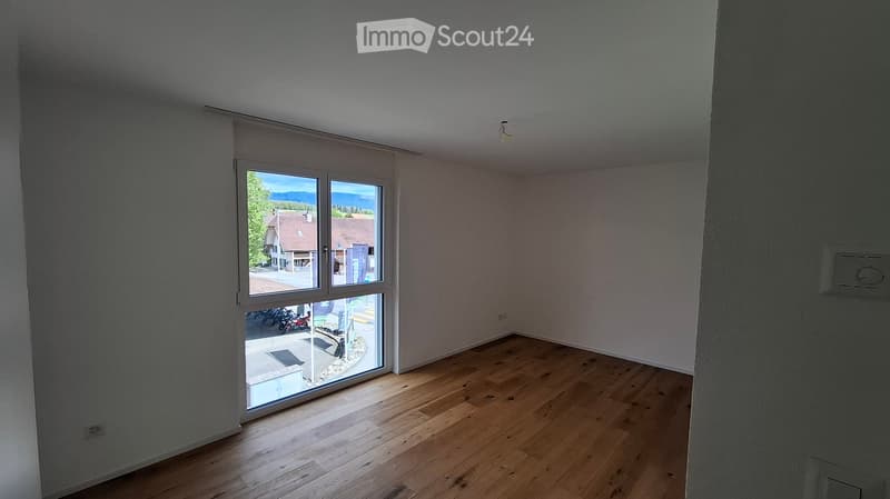 Empty room with wooden flooring and large windows offering a view of the street and surrounding houses.