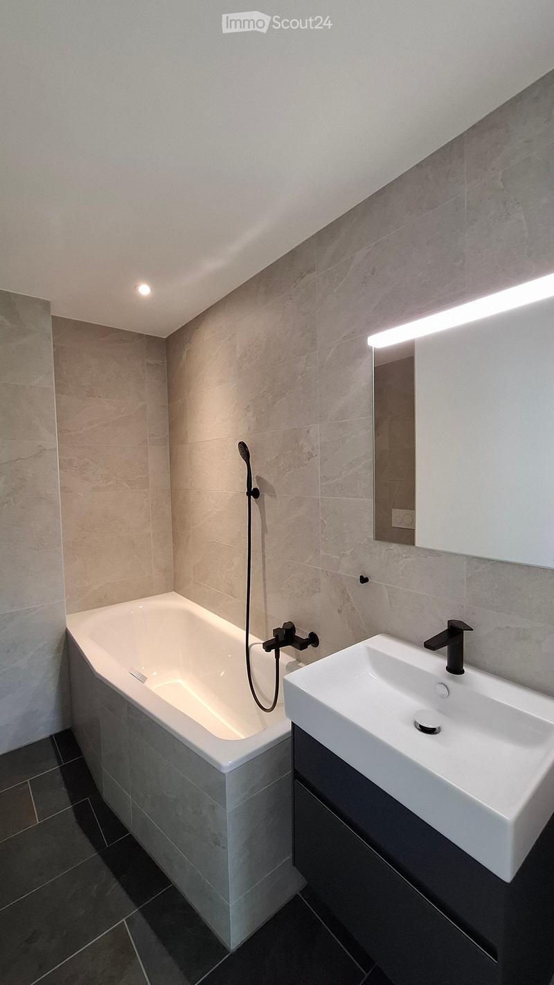 Modern bathroom with a white bathtub, a white sink, a black faucet, a mirror with built-in lighting, and a showerhead attached to the wall.