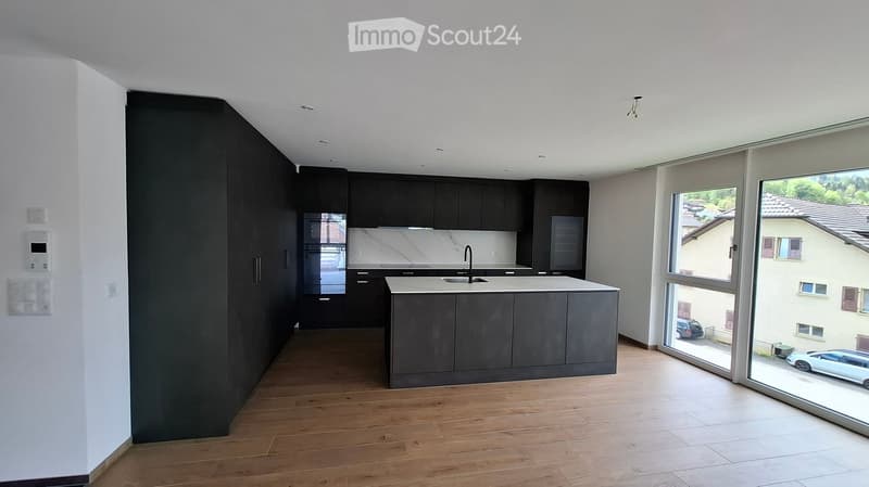 Modern kitchen with dark cabinets, a central island, and large windows overlooking a street with parked cars.