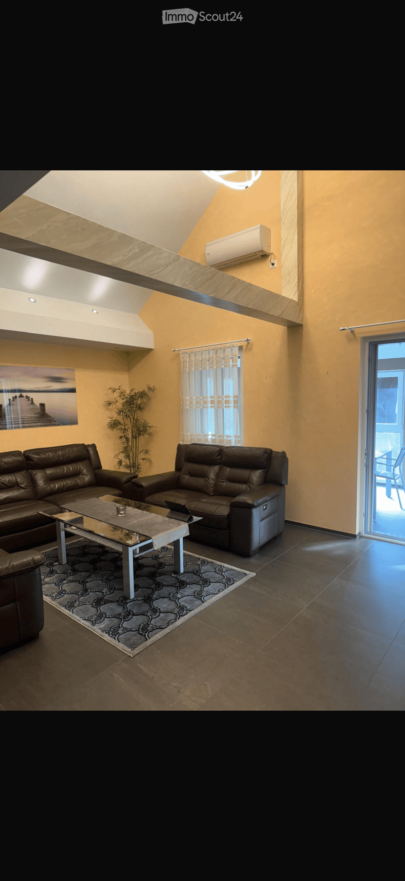 Modern living room with dark leather sofas, a glass coffee table, a patterned rug, and a large window with sheer curtains. An air conditioning unit is mounted on the wall, and a painting hangs on the wall.