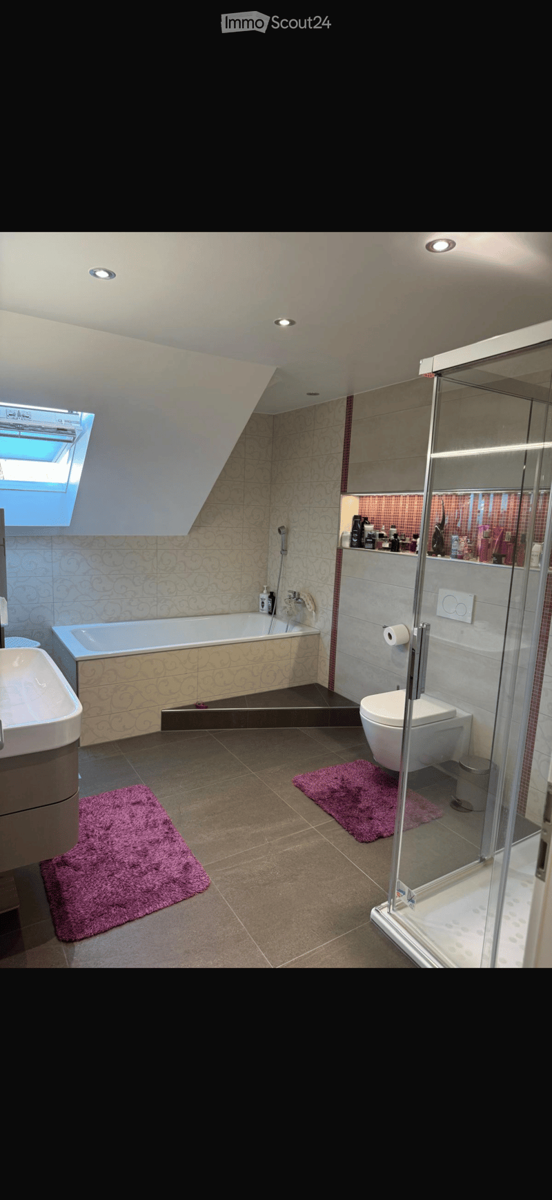 Modern bathroom with a bathtub, toilet, sink, and walk-in shower. Features decorative tiles, built-in storage, and two purple rugs.