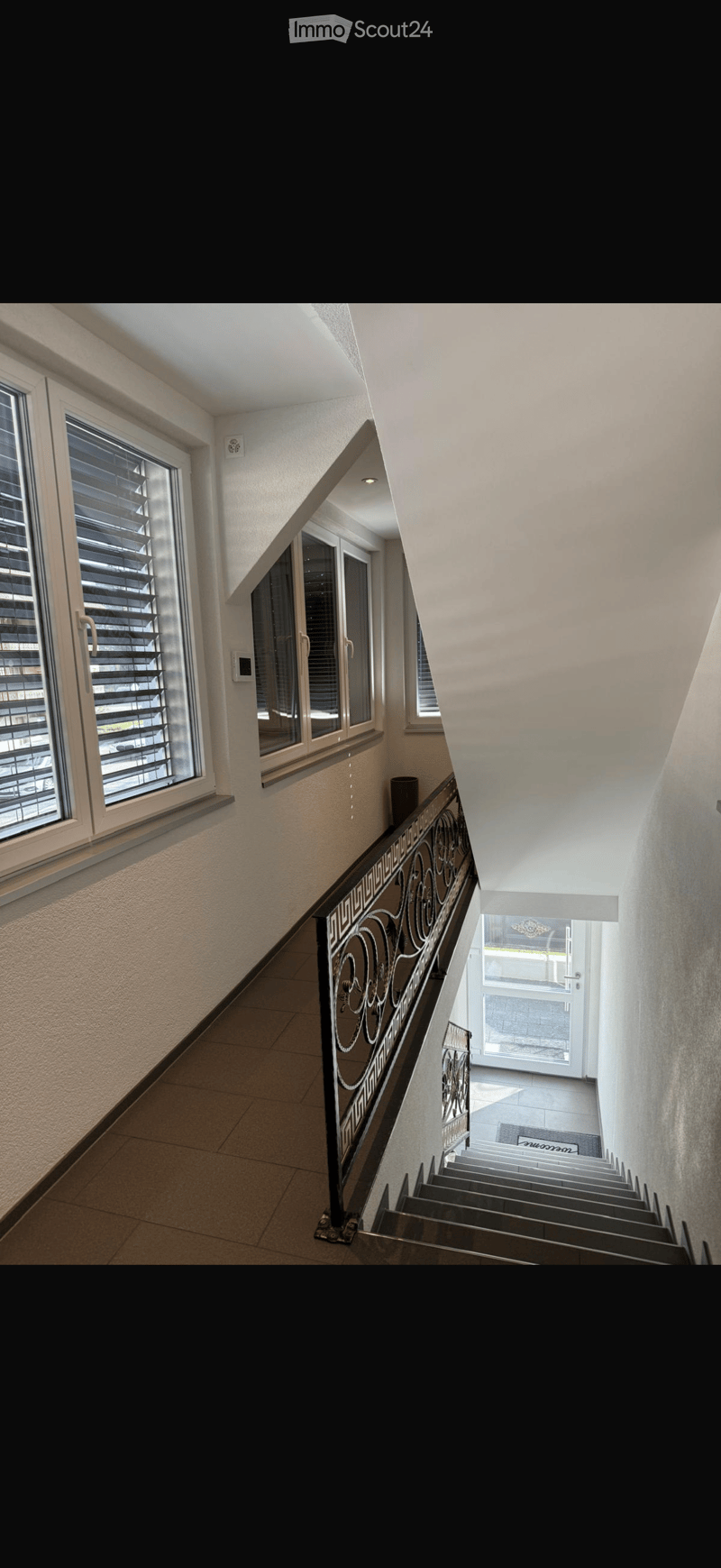 Spacious hallway with modern design, featuring a staircase with ornate metal railing, large windows with blinds, and a door leading to another area.