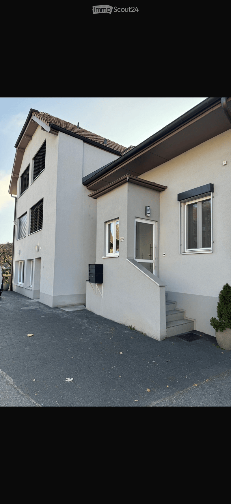 Two-story house with beige exterior, tiled roof, multiple windows, and a small staircase leading to the entrance.