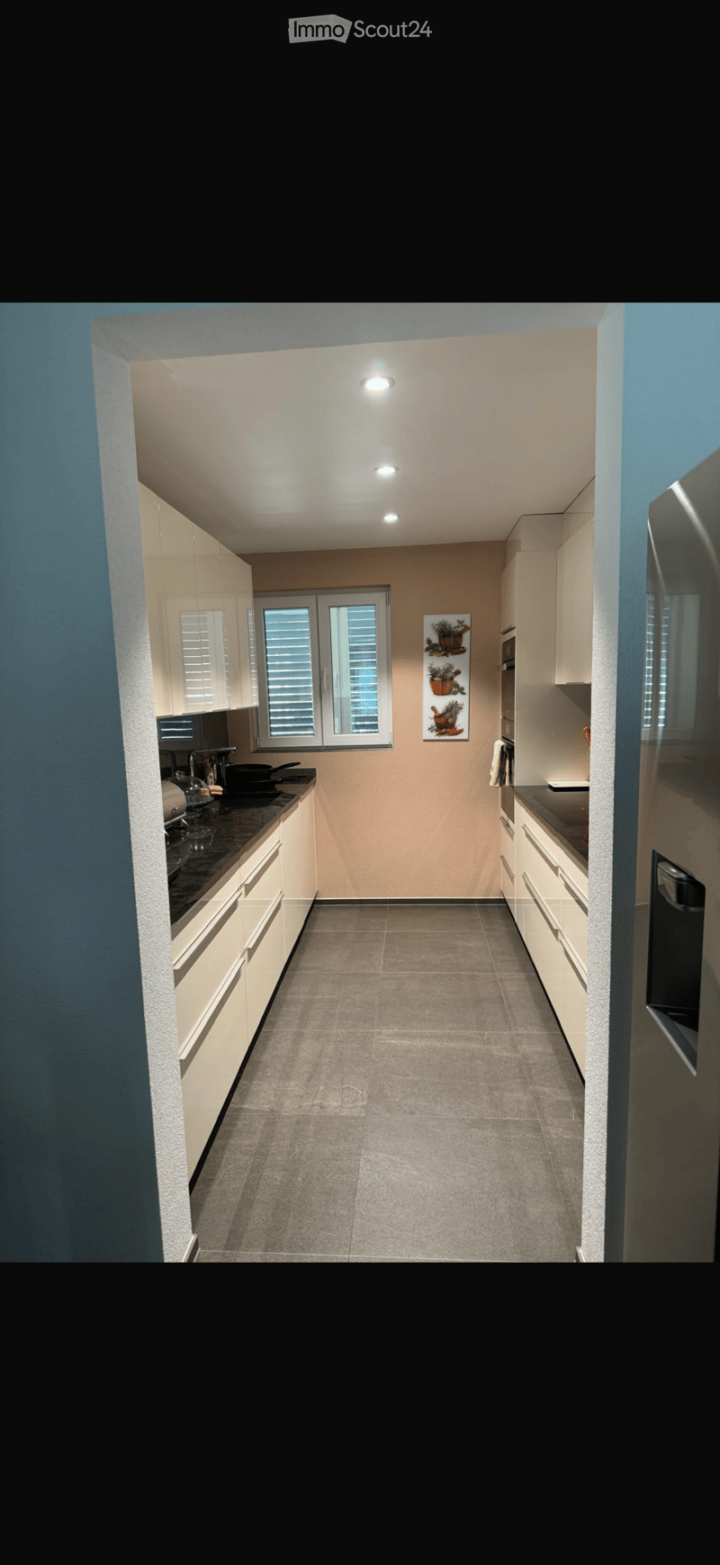 Modern kitchen with white cabinets, dark countertops, and a window with blinds. Includes a stove, sink, and refrigerator.