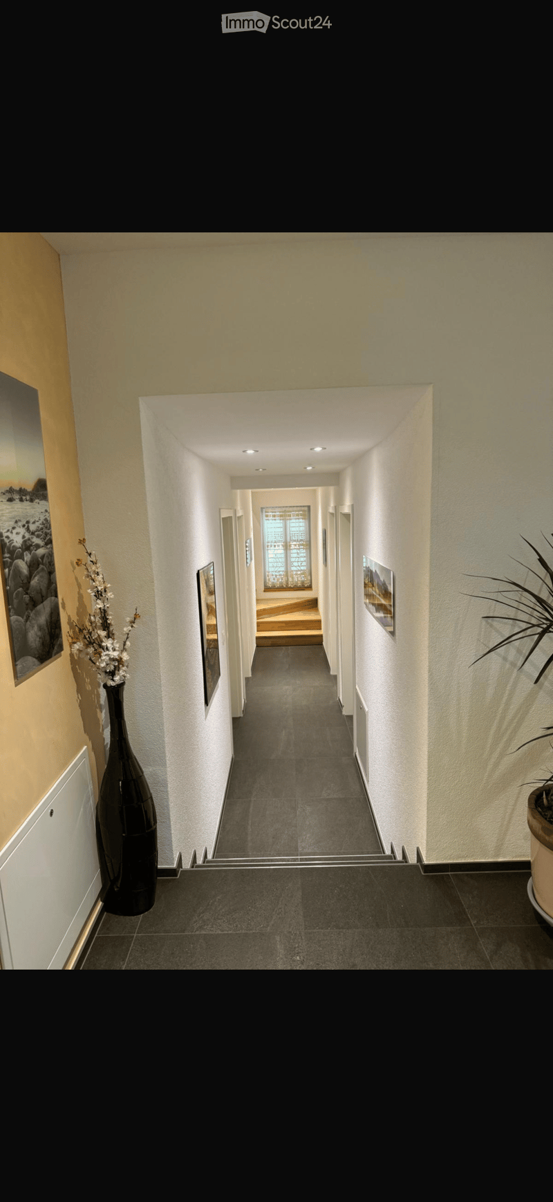 Modern hallway with grey tiled floor, white walls, recessed lighting, and framed artwork. Features a black vase with white flowers and a potted plant on the right side.