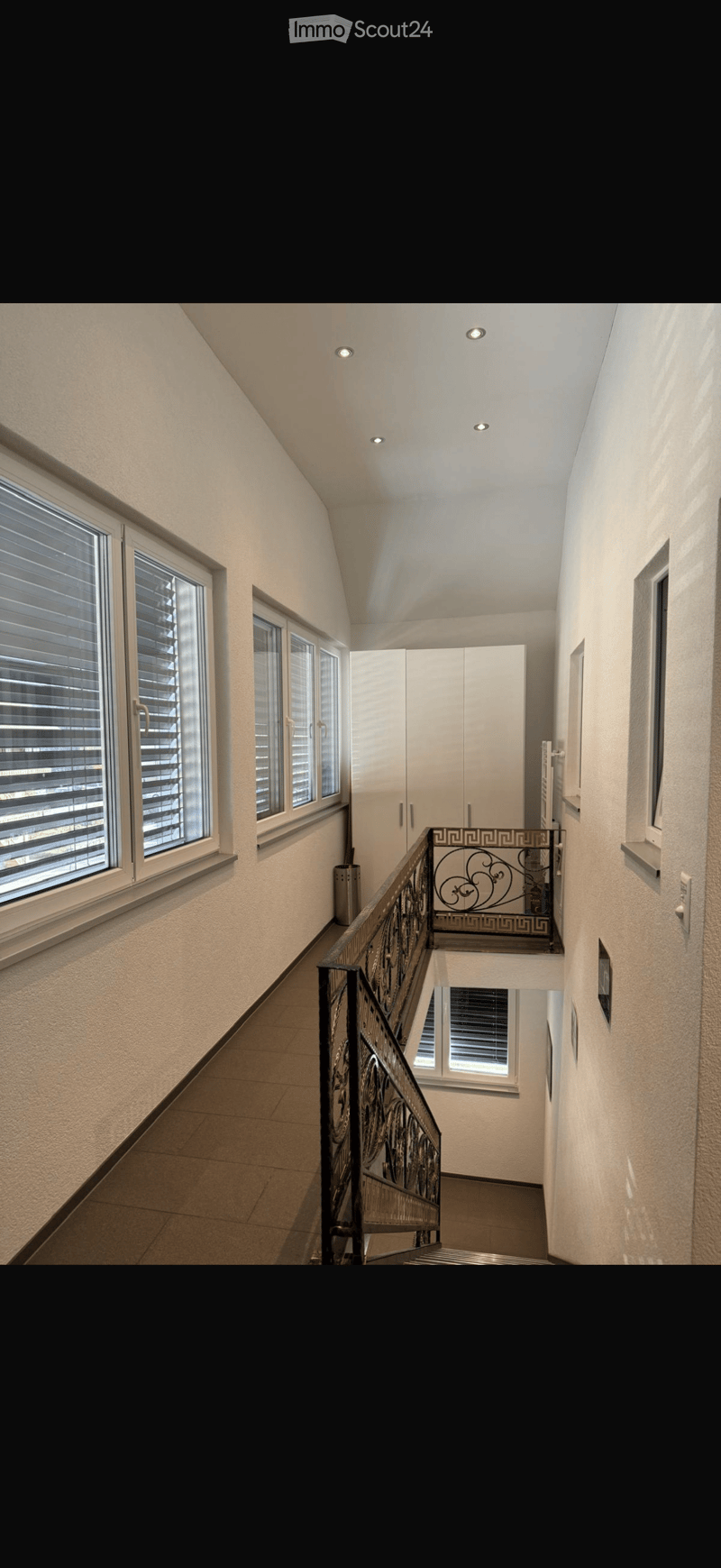 Spacious hallway with modern design, featuring recessed ceiling lights, large windows with blinds, and a decorative metal railing leading to a balcony.