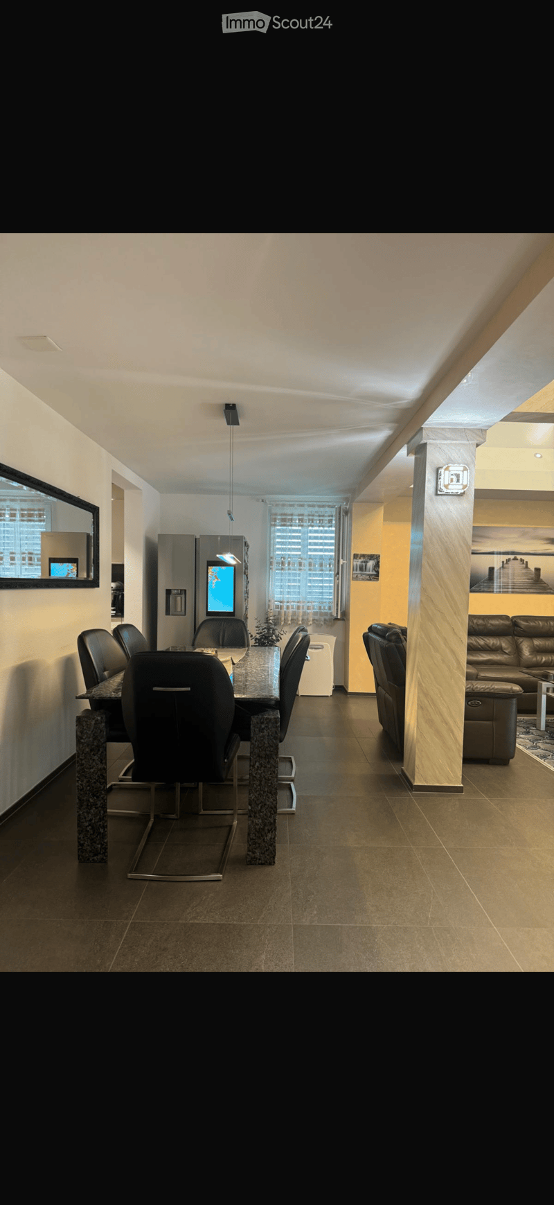 Modern open-plan living and dining area with a leather sofa, dining table with chairs, refrigerator, and a large screen on the wall.