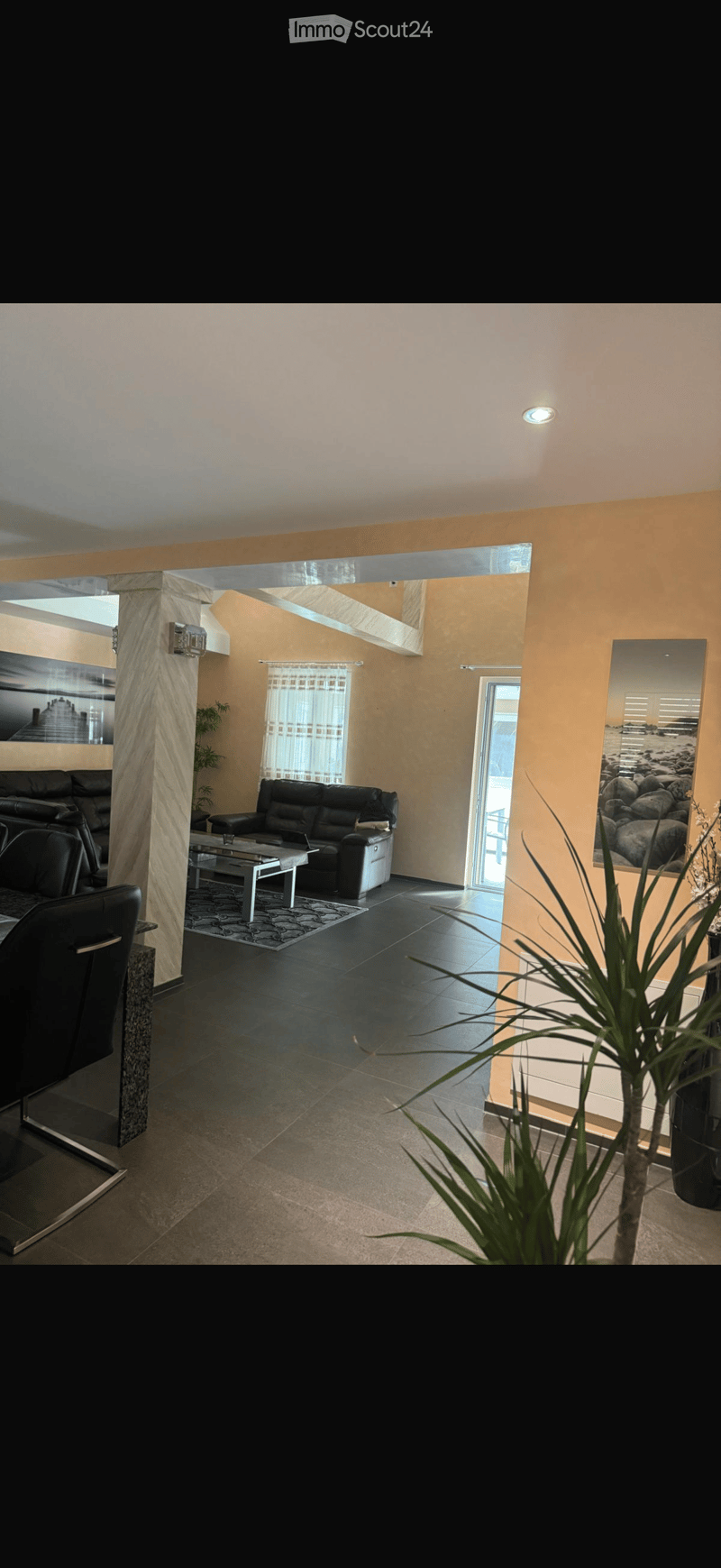 Modern living room with black leather sofa, coffee table, and large mirror. Features a high ceiling, large windows, and decorative plants.