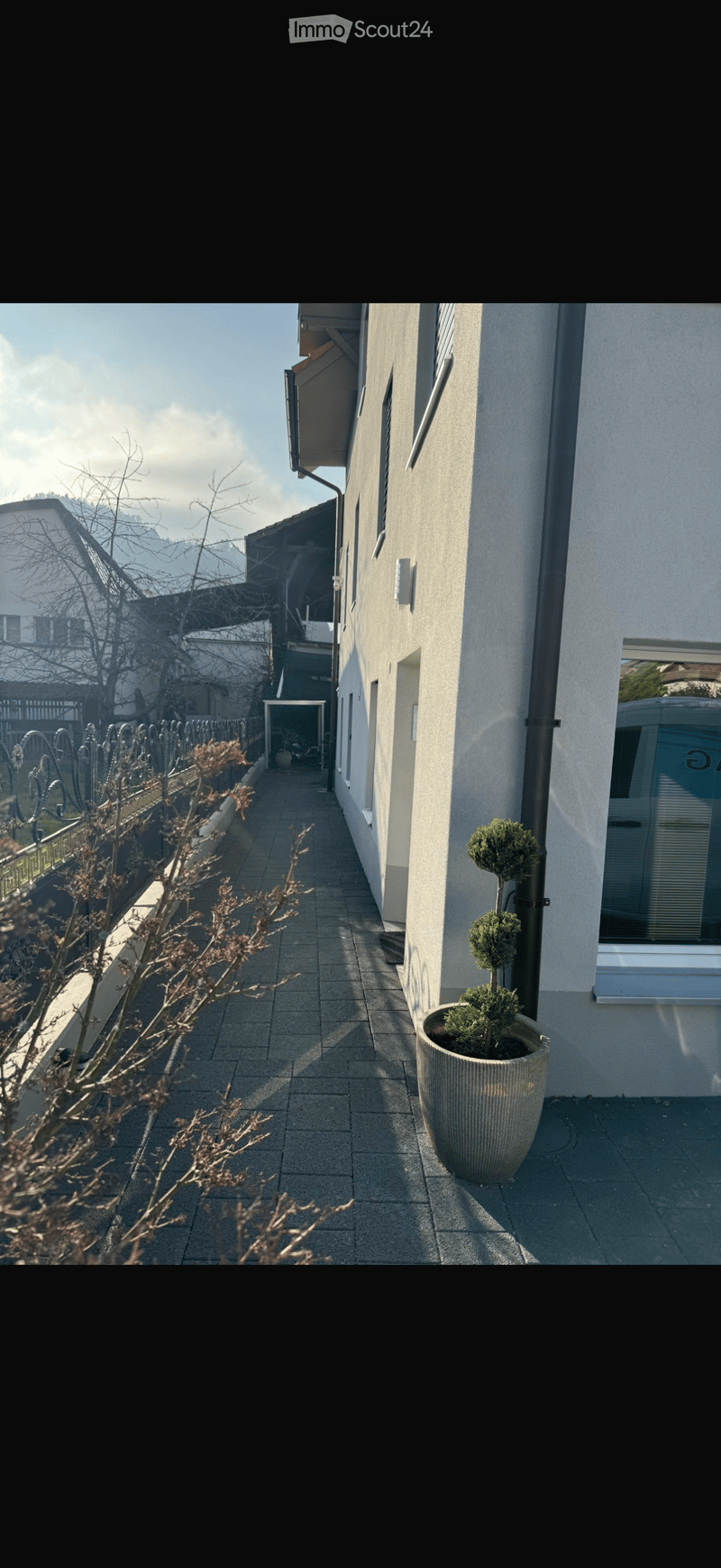Narrow paved pathway leading to a building entrance, flanked by a fence and potted plant, with a view of neighboring houses and trees.