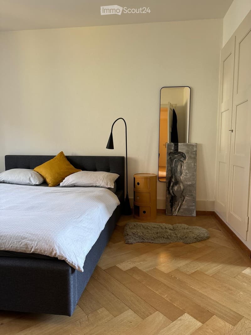 Modern bedroom with a dark grey bed, white bedding, yellow accent pillows, a floor lamp, a large mirror, a wooden nightstand, and a fur rug on the floor.