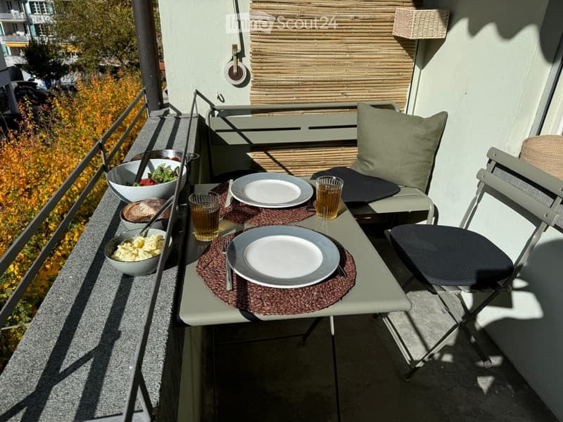 Balcony with a small table set for two, featuring plates, glasses, bowls of food, and a chair with a cushion. The balcony has a railing and a view of autumn foliage.