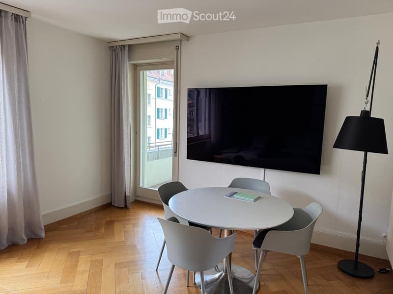 Modern dining area with a round white table, four grey chairs, a large flat-screen TV mounted on the wall, and a tall floor lamp with a black shade. The room features wooden flooring and large windows with grey curtains.