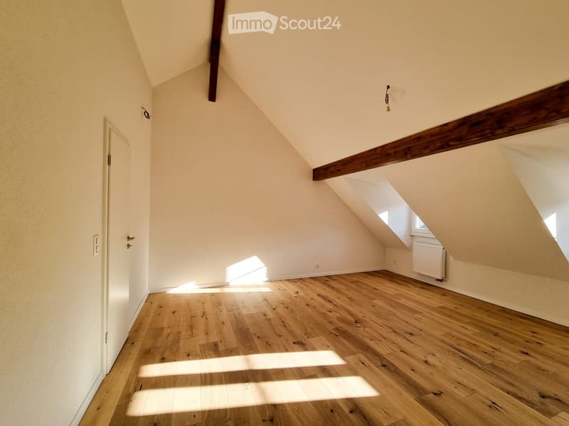 Empty attic room with wooden flooring, slanted ceilings, and exposed wooden beams. Natural light streams in through skylights, and there is a white door on the left side.