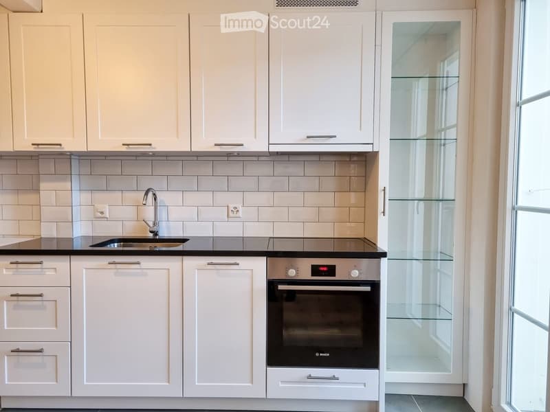 Modern kitchen with white cabinets, stainless steel sink, and Bosch oven. Features a tiled backsplash and a large window.
