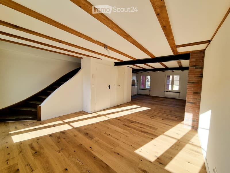 Empty room with wooden floor, exposed wooden beams on the ceiling, white walls, and a staircase leading to another level.