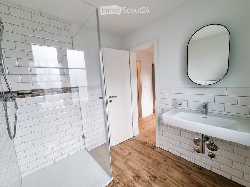 Modern bathroom with white tiled walls, a glass-enclosed shower, a white sink with a modern faucet, and a large round mirror.