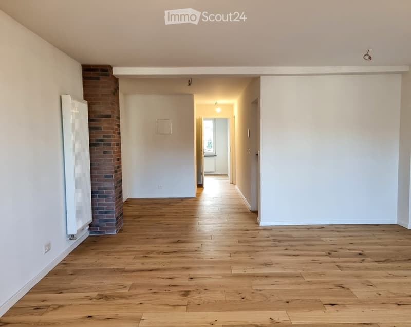 Empty hallway with wooden flooring, white walls, and a brick column on the left side. Natural light streams in from an open doorway at the end.