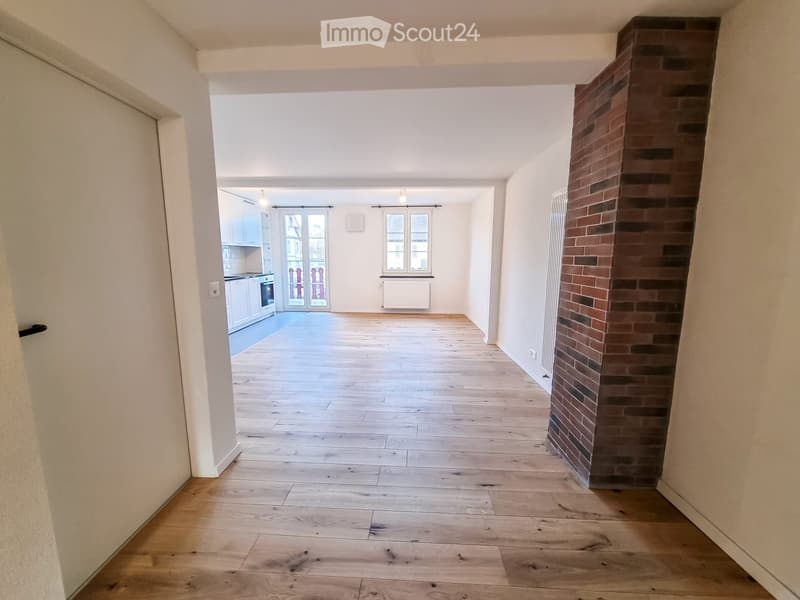 Empty room with wooden flooring, white walls, and a brick pillar. The room is connected to a kitchen area with white cabinets and a window.