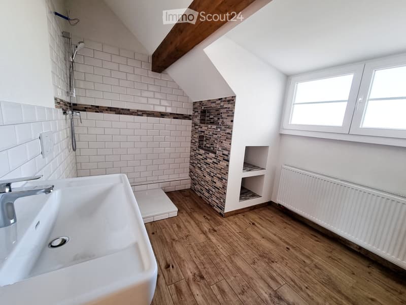 Modern bathroom with white tiles, wooden flooring, a large sink, a shower area, and a built-in shelf. The room features a slanted ceiling with exposed wooden beams and a window providing natural light.