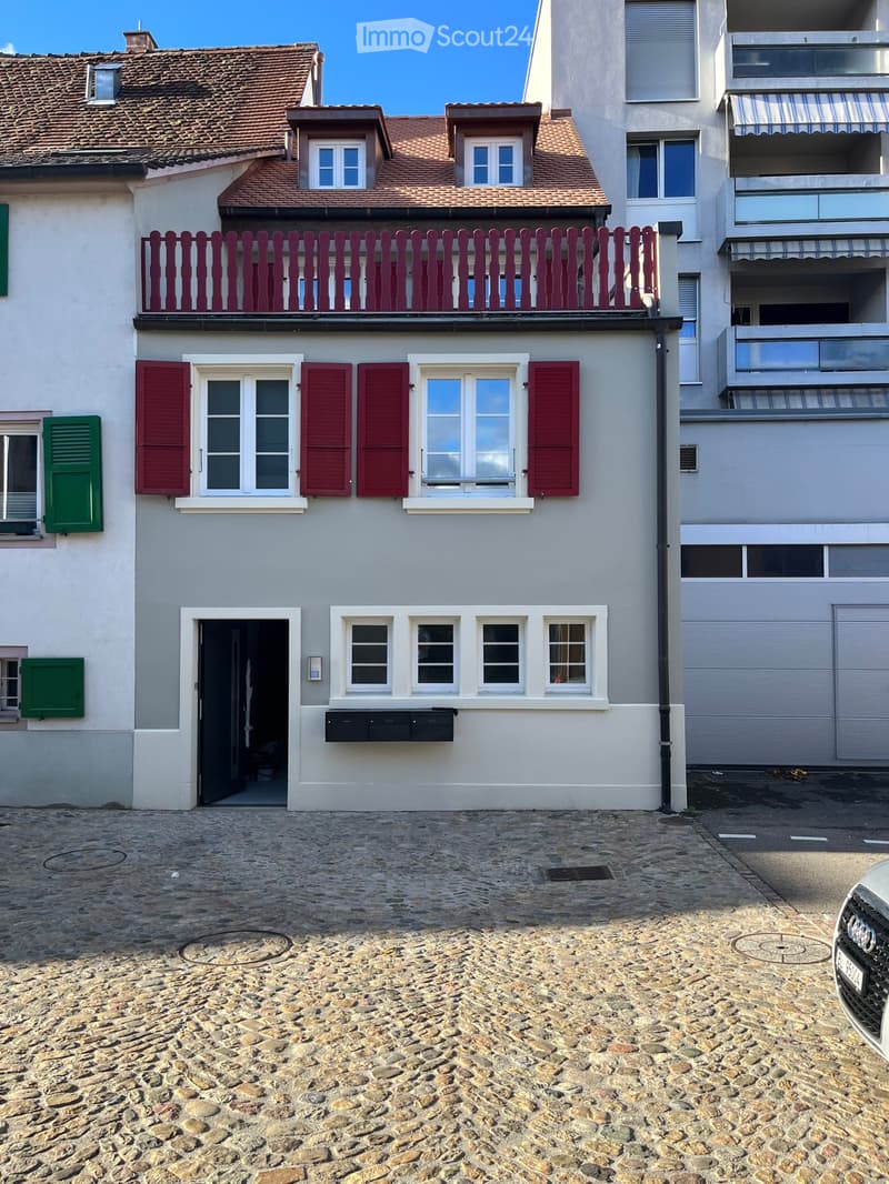 Two-story house with light gray walls, red shutters, and a red balcony railing. The ground floor has a black door and several windows, while the upper floor features additional windows and a small balcony. The roof is tiled with brown tiles and has dormer