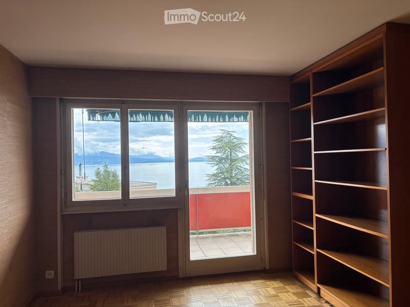 Empty room with wooden flooring, built-in wooden shelving unit, large window with scenic lake view, radiator, and sliding glass door leading to a balcony.
