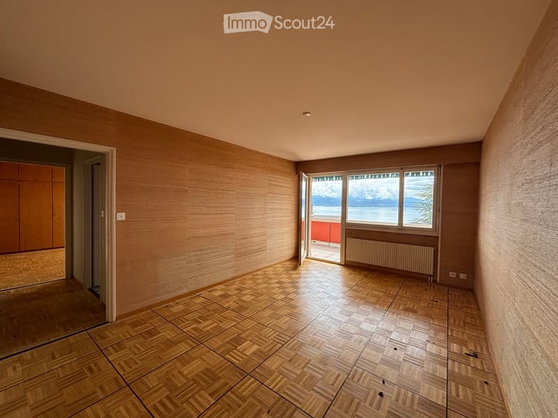 Empty room with wooden herringbone flooring, light wooden walls, large windows with a scenic view, and a radiator under the windows.
