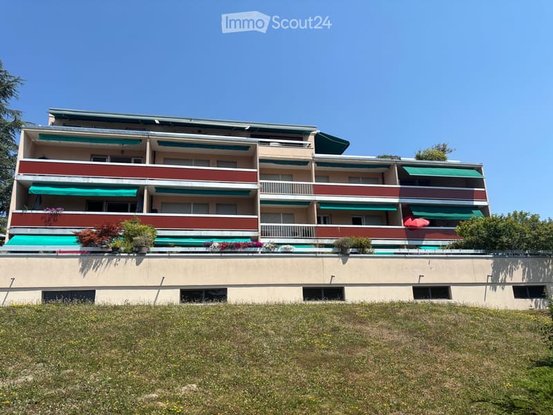 Multi-story residential building with balconies, green awnings, and potted plants on the lower level.