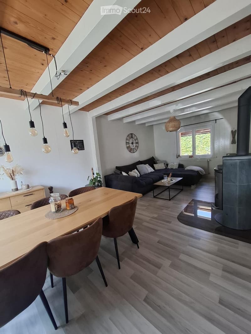 Modern living and dining area with exposed wooden beams, pendant lights, a dark grey sofa, a wooden dining table with chairs, and a stone fireplace.