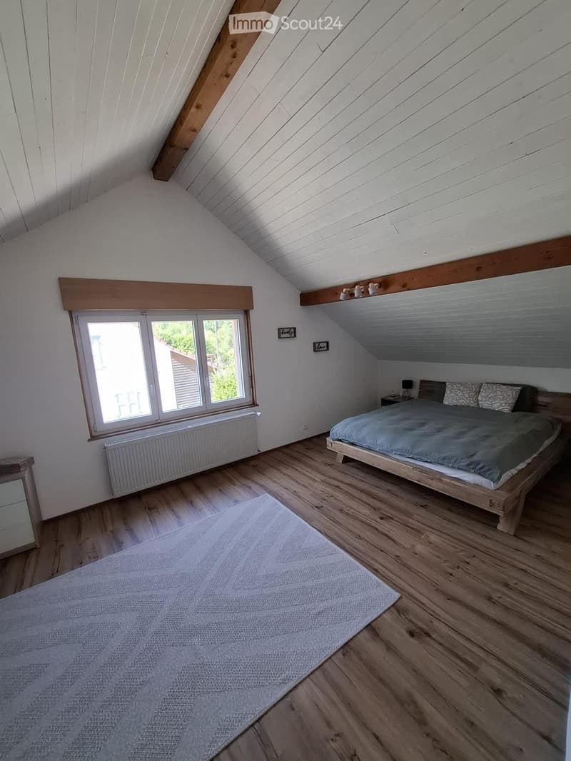 Cozy attic bedroom with wooden floors, slanted ceilings, large window, bed, nightstand, and lamp.