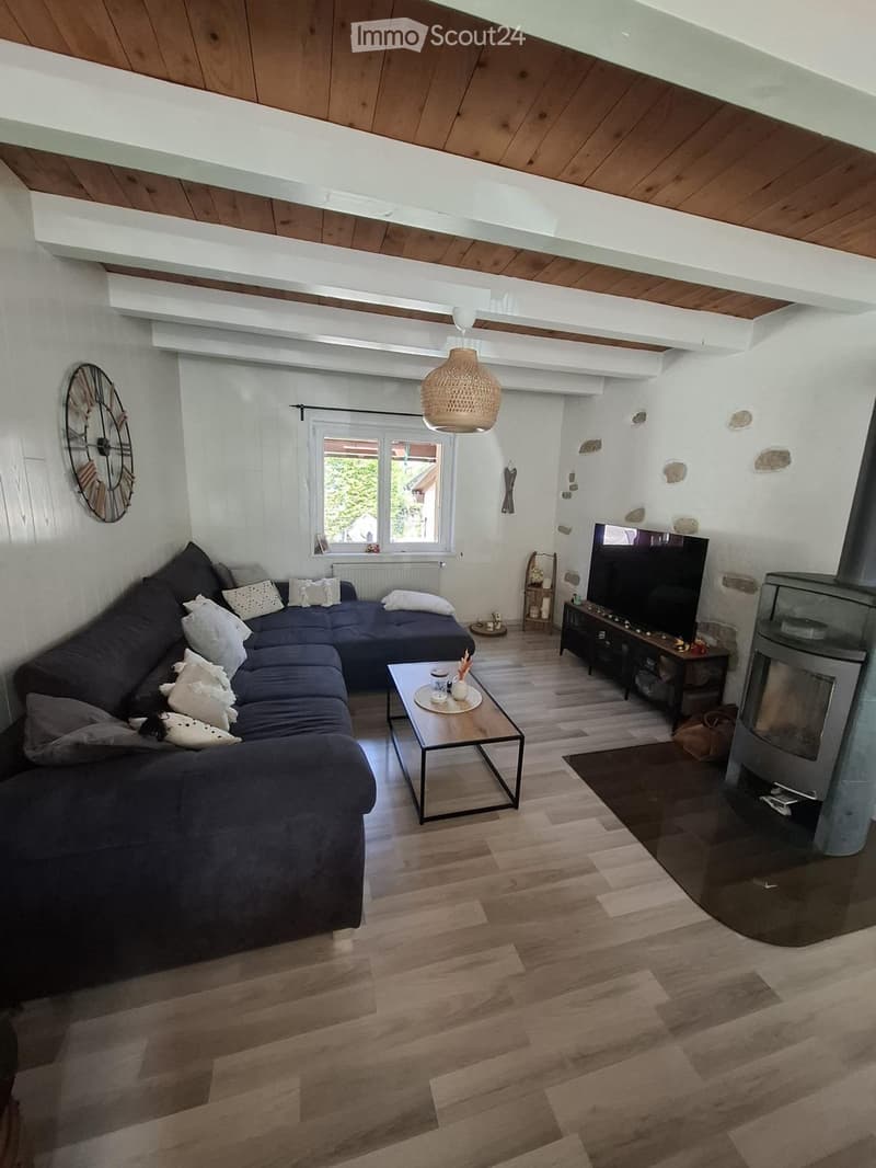 Modern living room with wooden flooring, a dark blue sectional sofa, a glass coffee table, a television, and a fireplace. The room features exposed wooden beams on the ceiling and a large window letting in natural light.