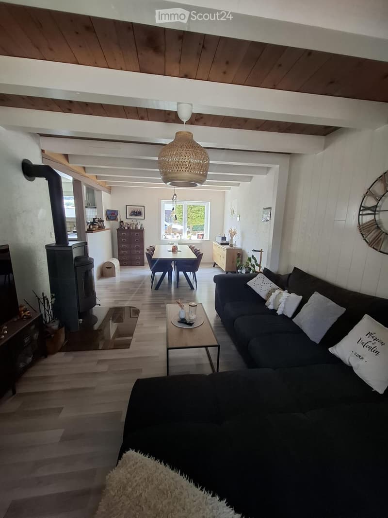 Open-concept living and dining area with exposed wooden beams, a dark grey sofa, coffee table, fireplace, dining table with chairs, and a hanging woven lamp.