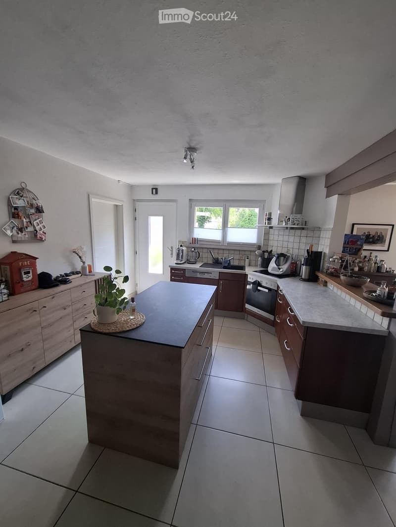 Modern kitchen with dark wooden cabinets, a central island, built-in appliances including an oven, dishwasher, and coffee maker, and a potted plant on the island.