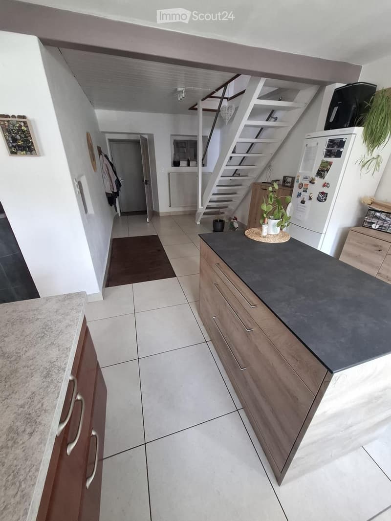 Modern kitchen with wooden cabinets, dark countertop, white tiled floor, and a small plant on the counter. A staircase leads to an upper level.