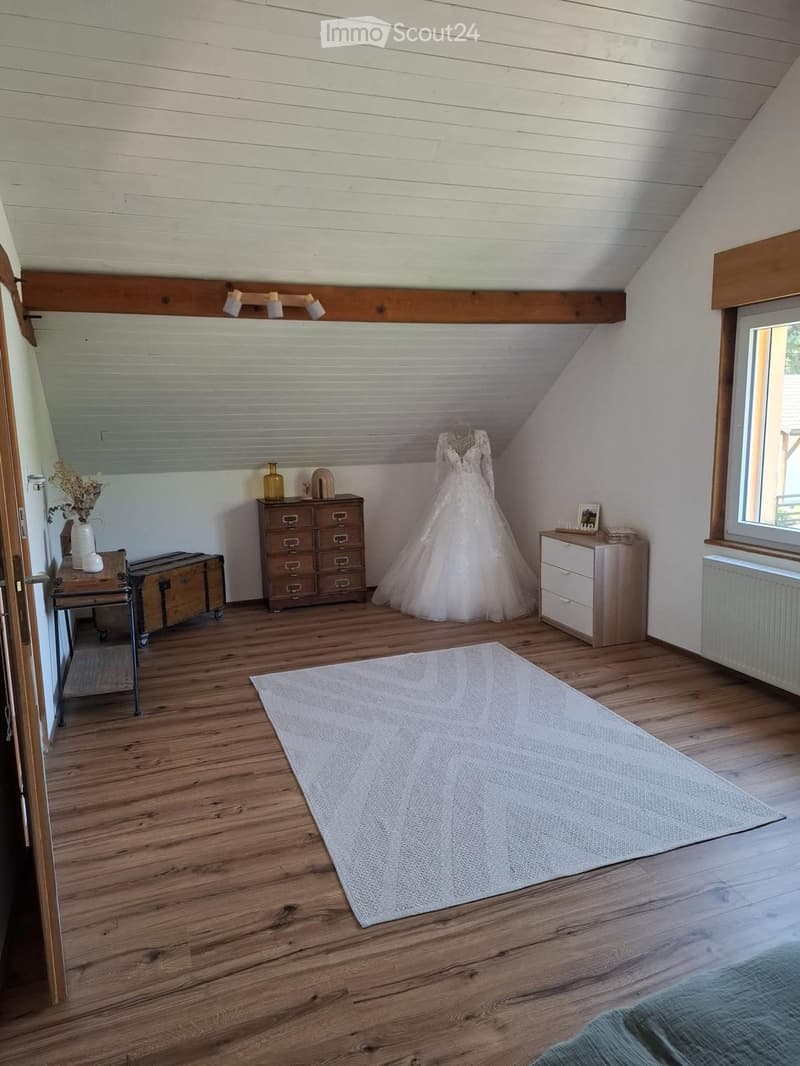 A cozy attic room with wooden floors, slanted ceilings, and exposed wooden beams. Features include a wedding dress displayed on a mannequin, a chest of drawers, a small table with a vase, and a large window letting in natural light.