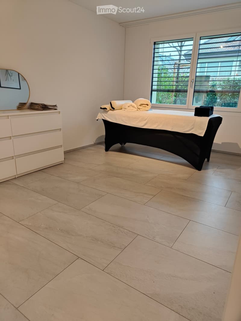 Bathroom with a massage table, a mirror, and a towel rack. The room features light-colored tiled flooring and white walls.