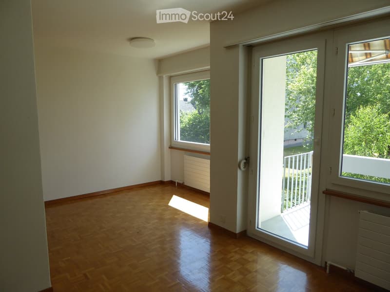 Empty room with wooden flooring, white walls, large windows, and a sliding glass door leading to a balcony.