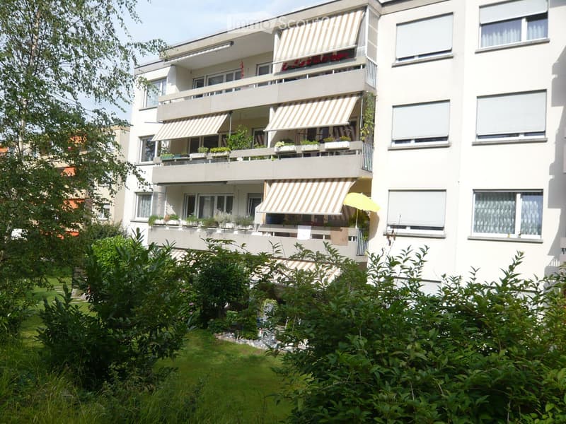 Modern multi-story apartment building with balconies, striped awnings, and potted plants. Greenery surrounds the building.