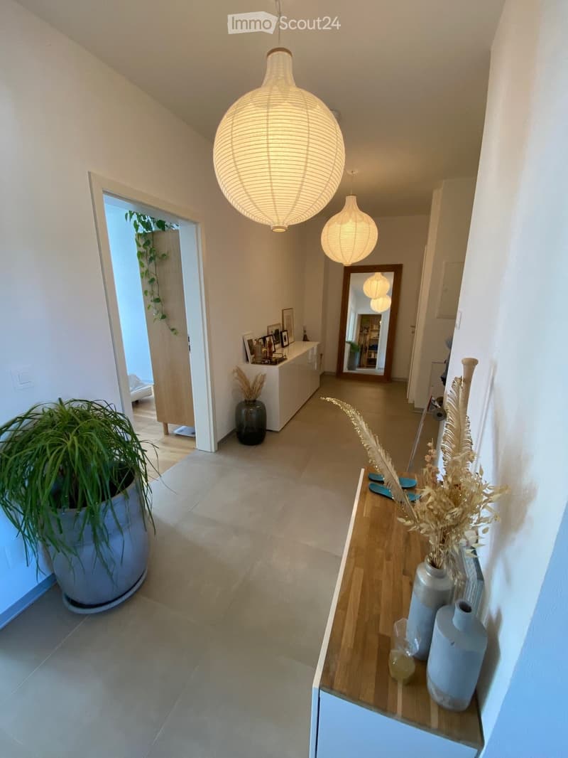 wide hallway, beige tiled floor, lamps, wooden console table with vases, mirror, tall potted plant, hallway leading to another room