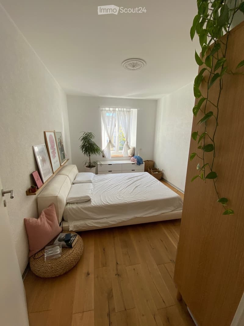 A cozy bedroom with a double bed, wooden floor, a shelf with framed pictures, a potted plant, a cabinet with drawers, and a window with white curtains.