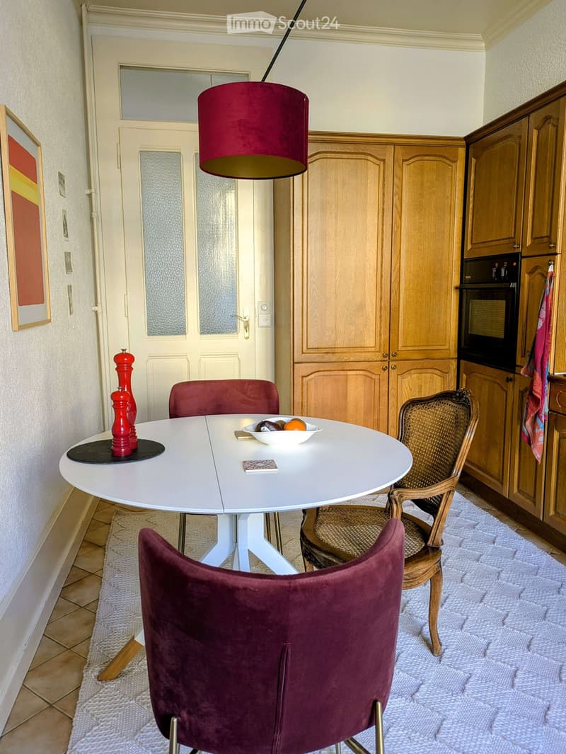 A cozy kitchen with a round white dining table, red chairs, a wicker chair, a bowl of fruit, a pepper grinder, wooden cabinets, and an oven.
