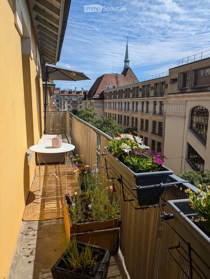 Balcony with a small table, chair, and multiple flower pots, overlooking a cityscape with historic buildings and a spire.