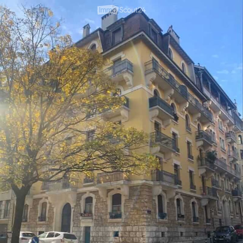 A multi-story residential building with yellow facade, multiple balconies, and ornate architectural details. The building has a stone base and is surrounded by trees and parked cars.
